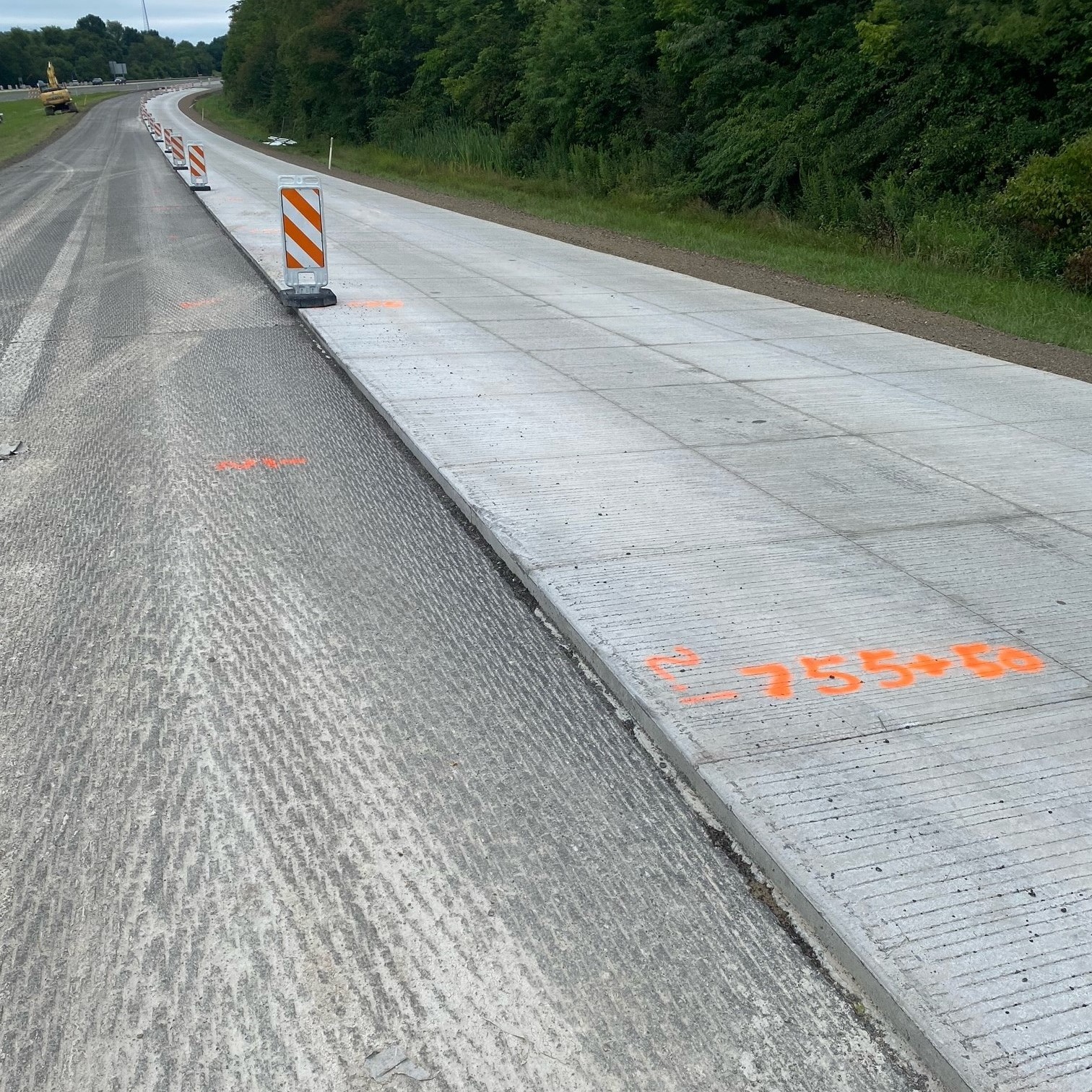 An image of a tree-lined, four-lane roadway with a grassy median between either side of the roadway with a line of orange and white work zone barricades between the completed lane and the lane be prepared for the concrete overlay